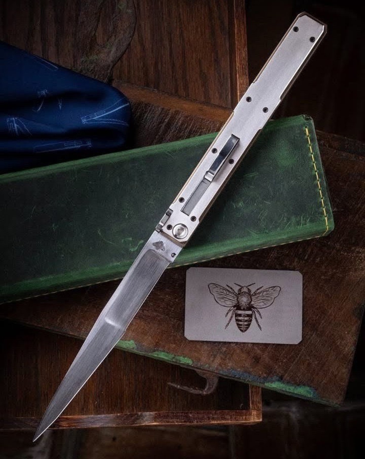 Silver folding knife on green leather beside an engraved bee card